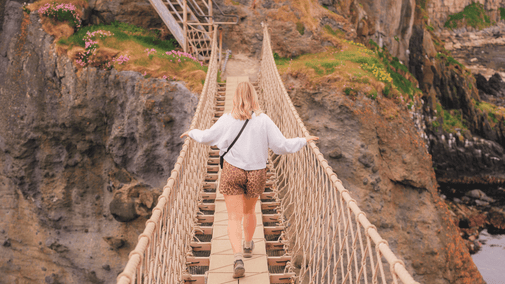 A visitor wearing a grey jumper and leopard print shorts, walking across Carrick-a-Rede Rope Bridge, captured from the island